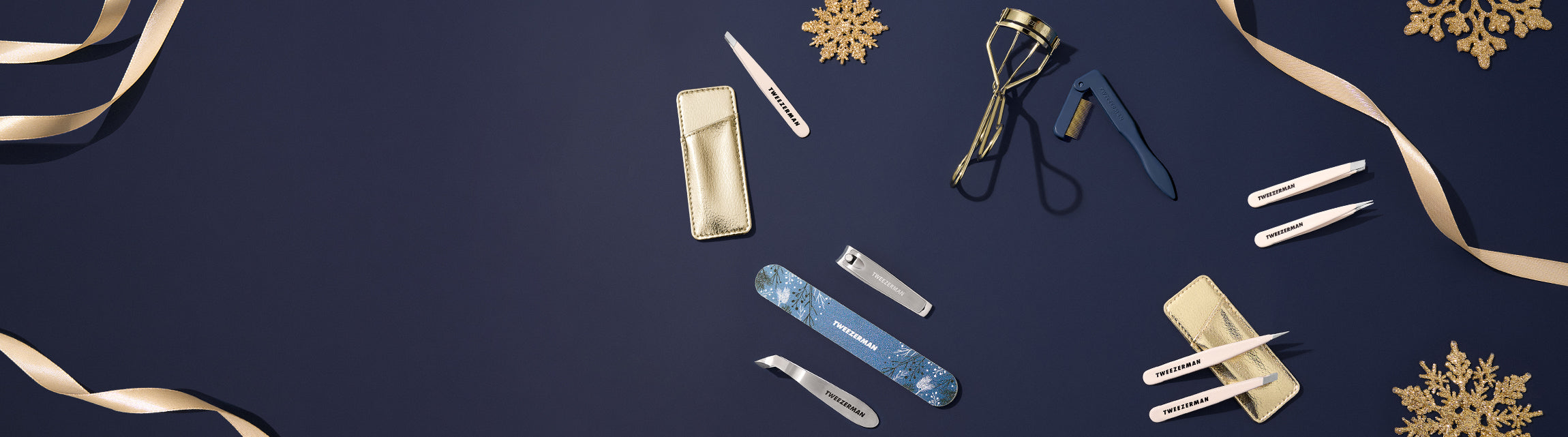 A flat-lay arrangement of various beauty and skincare tools on a dark navy blue background with gold ribbons and snowflake decorations. The tools include a blue nail file, a beige makeup brush, a black eyelash curler, and several silver metal spatulas and applicators.