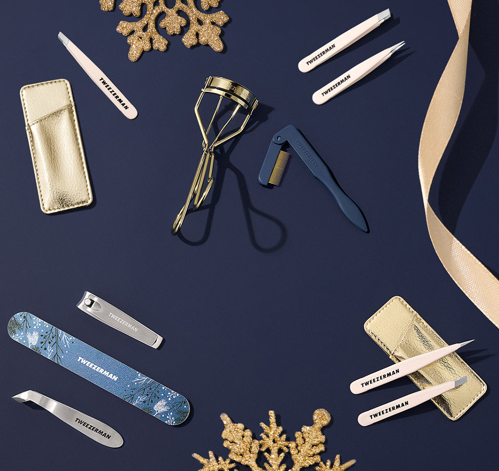 A flat-lay arrangement of Tweezerman nail and beauty tools on a dark navy background, including tweezers, a metal eyelash curler, a blue nail file, metal cuticle tools, and metallic pouches, with decorative gold snowflakes and a beige ribbon.
