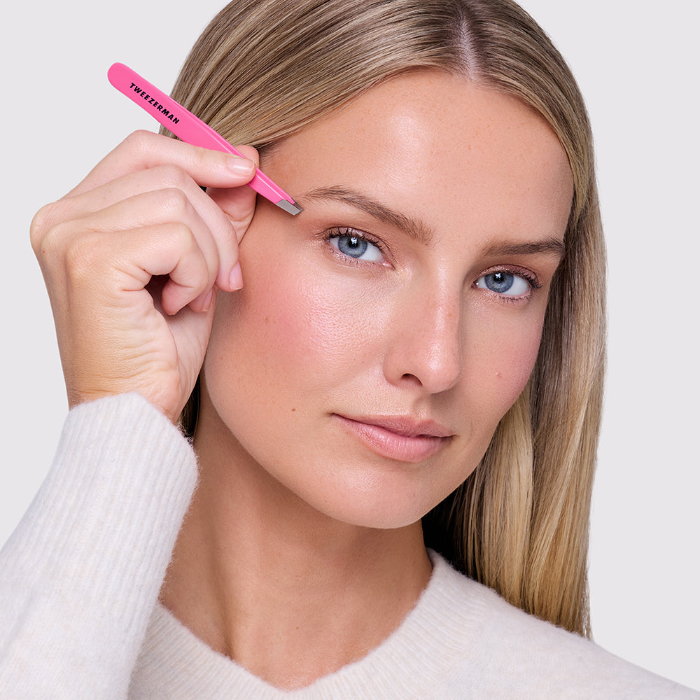 A close-up of a woman holding a pink Tweezerman slant tweezers tool near her eyebrow, wearing a white knit sweater against a light gray background.