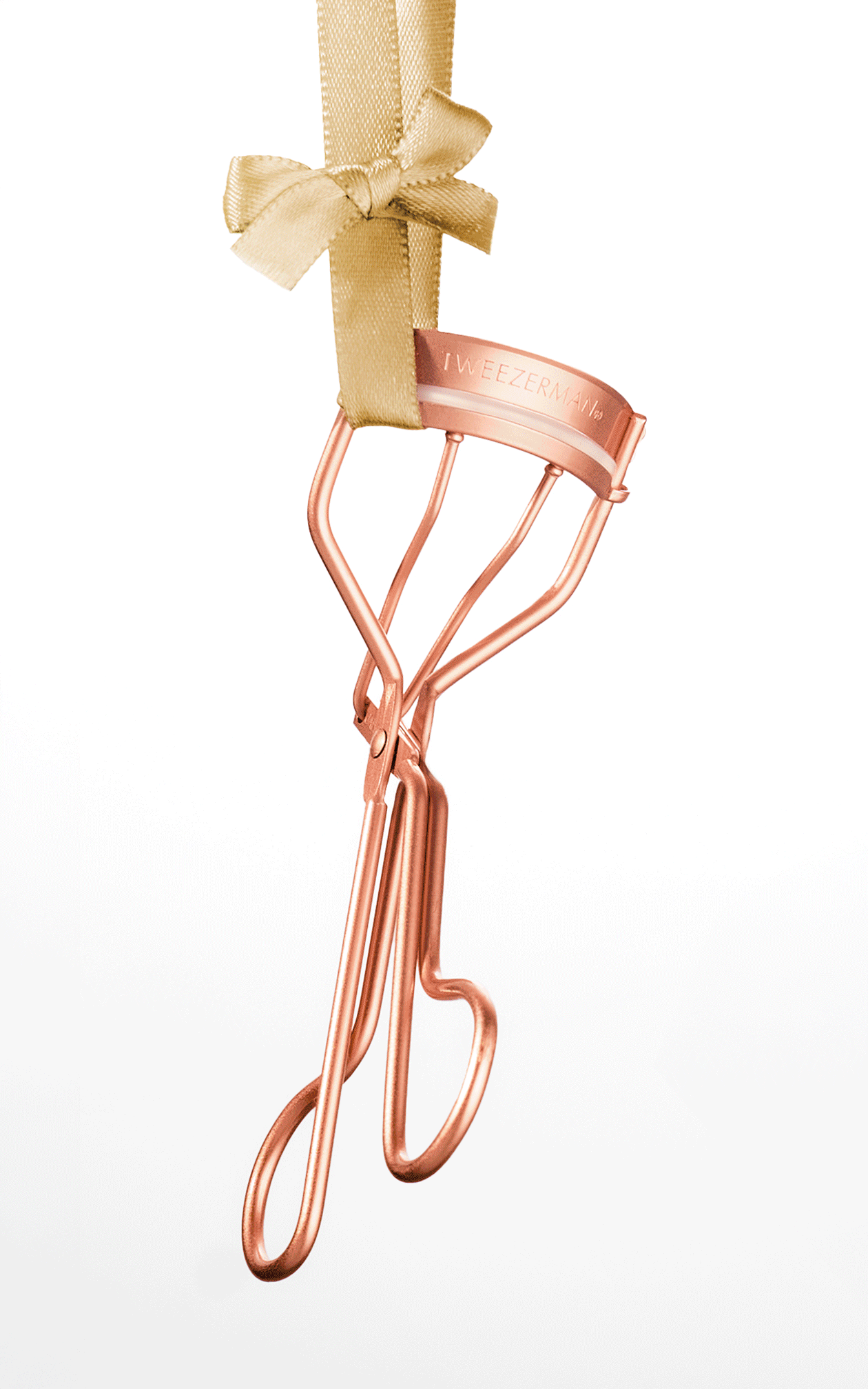 A set of rose gold beauty tools hanging from gold ribbon against a white background, including an eyelash curler, brow scissors, a brow spoolie, and a mini slant tweezer