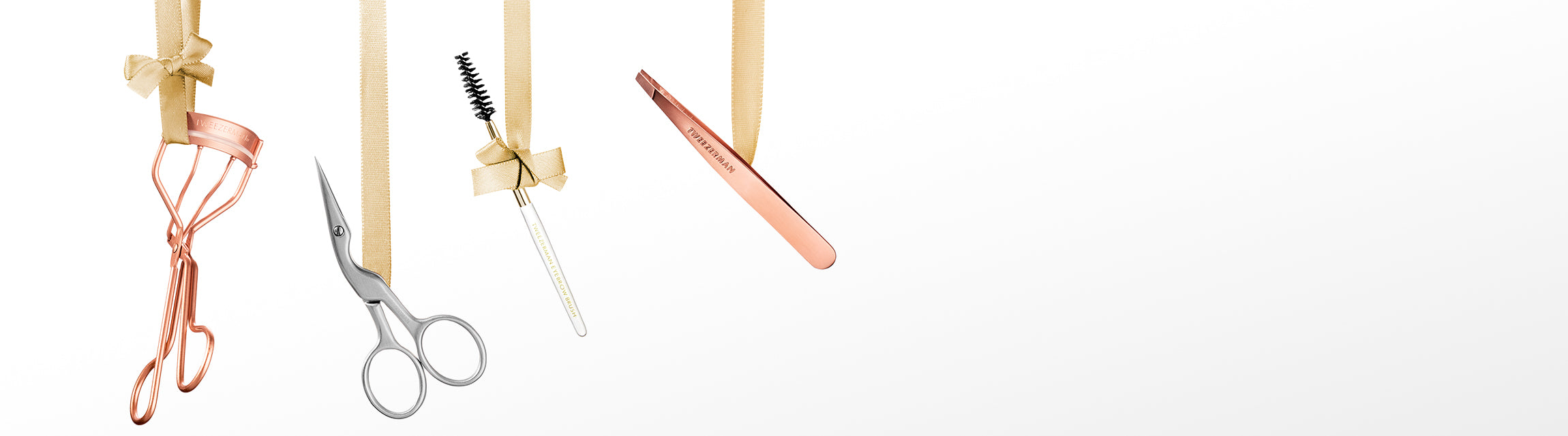 A set of rose gold beauty tools hanging from gold ribbon against a white background, including an eyelash curler, brow scissors, a brow spoolie, and a mini slant tweezer