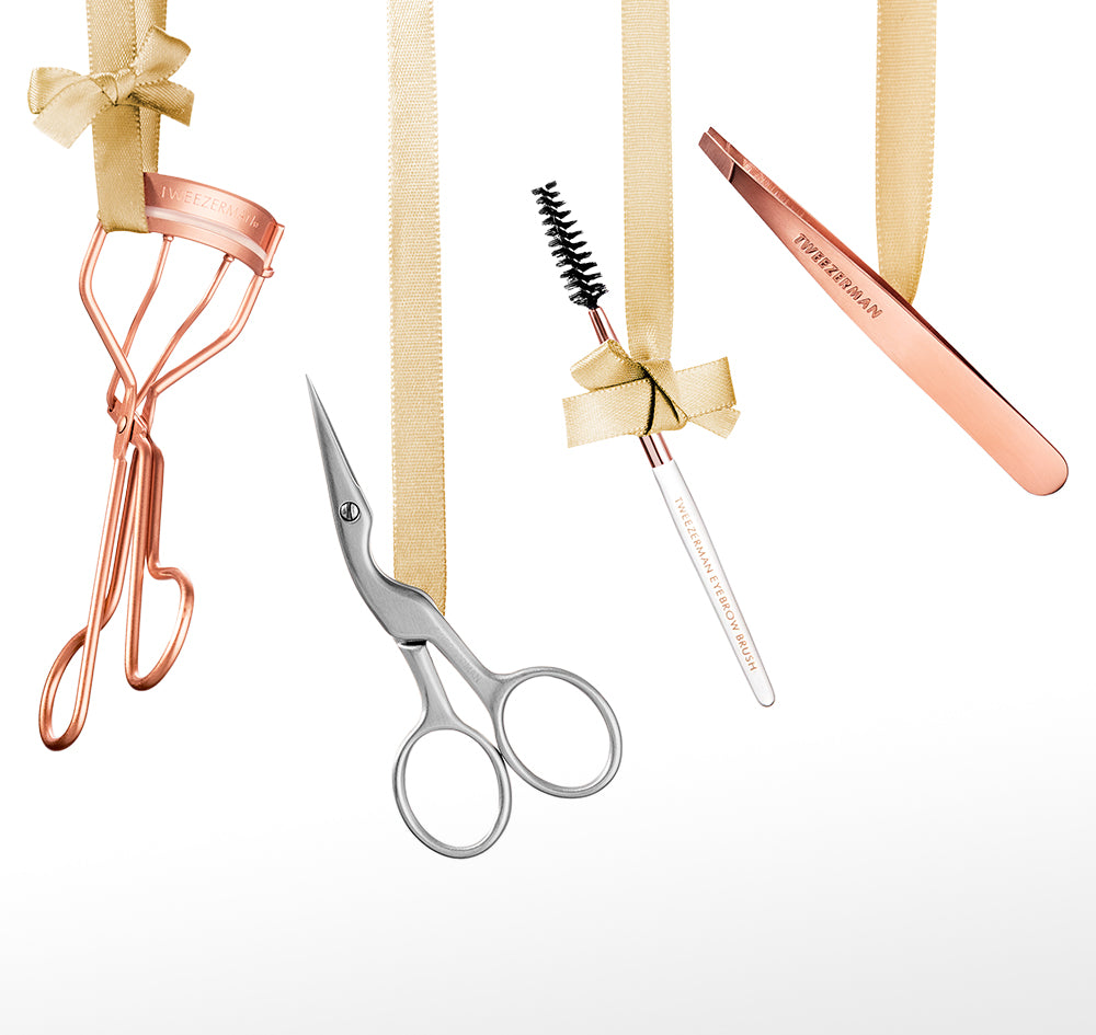 A set of rose gold beauty tools hanging from gold ribbon against a white background, including an eyelash curler, brow scissors, a brow spoolie, and a mini slant tweezer