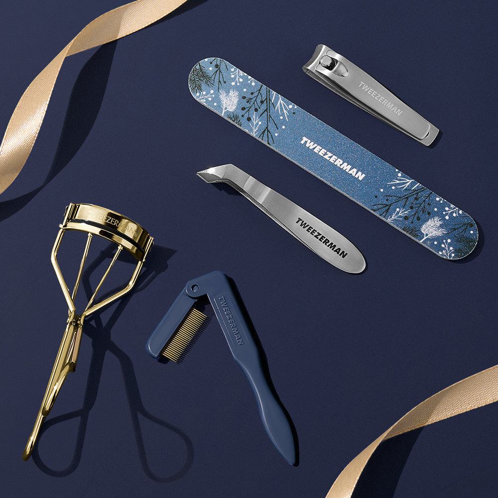 A set of tweezman eyebrow and eyelash tools from the latest Evening Glow collection arranged on a dark navy background with gold ribbon accents. The set includes a gold eyelash curler, a blue floral nail file, stainless steel mini cuticle nail clipper, and a blue eyebrow comb.