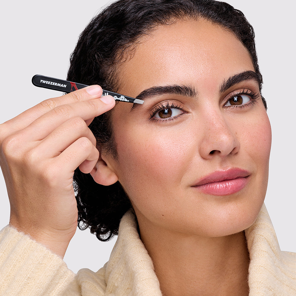 A close-up of a woman using a  black Tweezerman slant tweezer from The Devil Wears Prada 2 Collection. held in her hand, wearing a cream-colored knit robe against a light gray background.