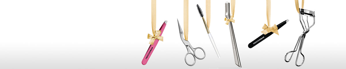 A row of beauty tools hanging from gold ribbons against a white background, including a pink Tweezerman slant tweezer, stainless steel brow scissors and spoolie. Brow razor a black slant tweezer, and a stainless-steel classic eyelash curler.