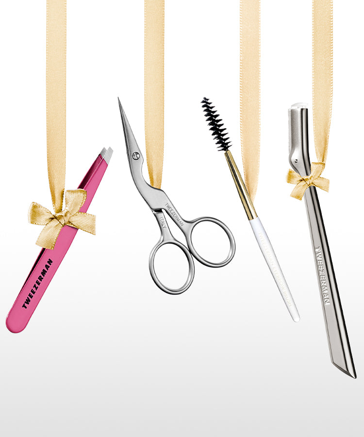 A row of beauty tools hanging from gold ribbons against a white background, including a pink Tweezerman slant tweezer, stainless steel brow scissors and spoolie. Brow razor a black slant tweezer, and a stainless-steel classic eyelash curler.