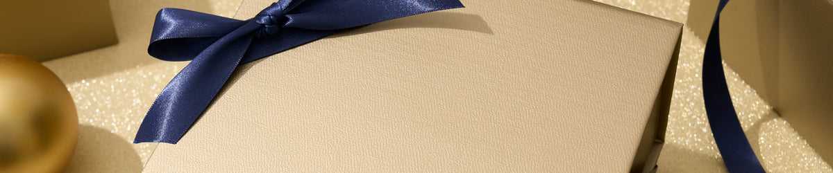 A close-up of a textured beige gift box tied with a wide navy blue satin ribbon bow, set against a glittery gold background.